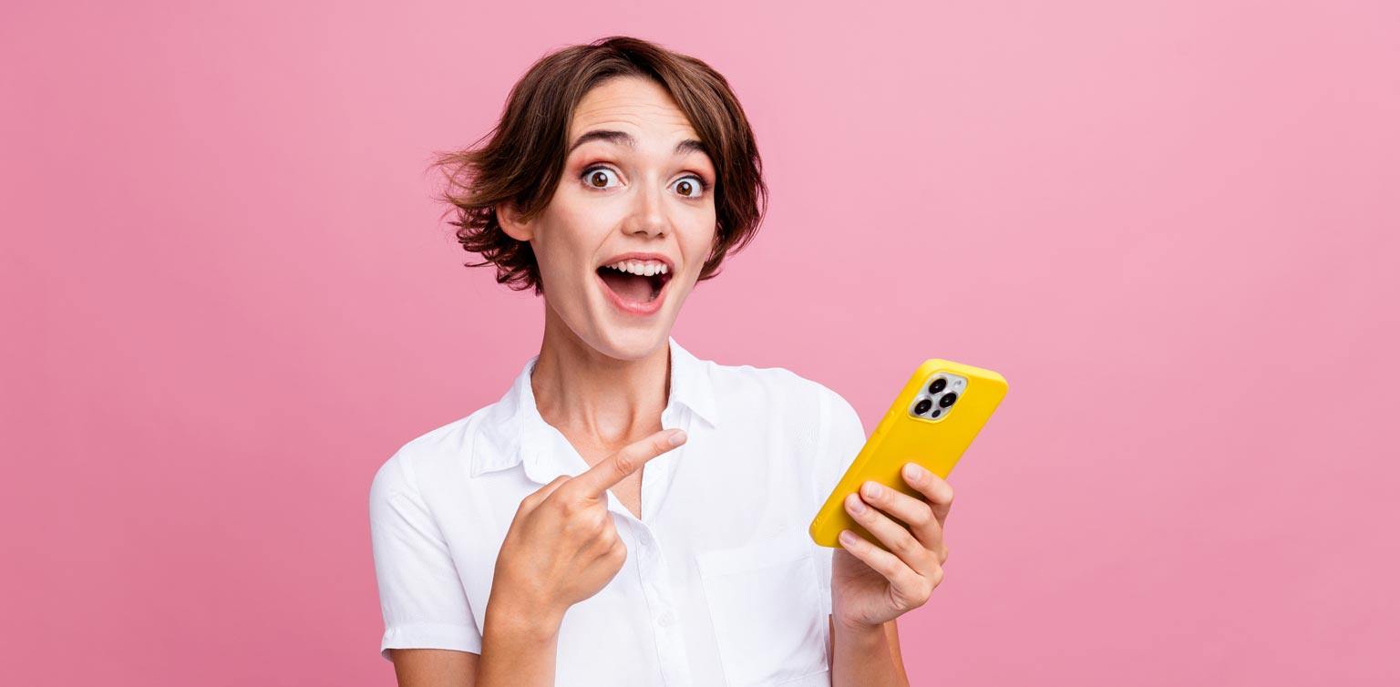 A photo of a woman pointing at her smart phone with an amazed look on her face.