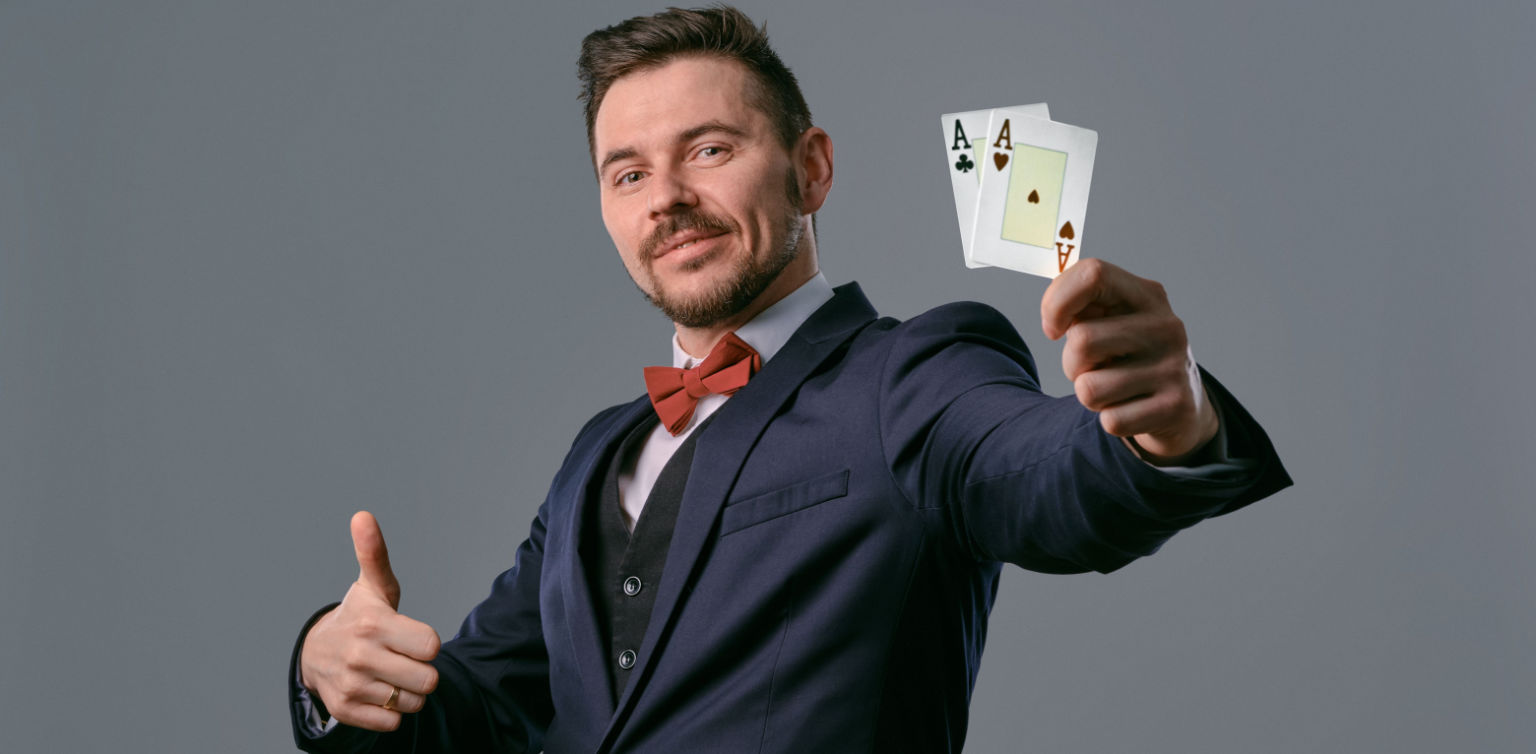 A photo of a smiling man in a suit holding a pair of Aces in one hand, giving a thumbs up with the other, on a grey background