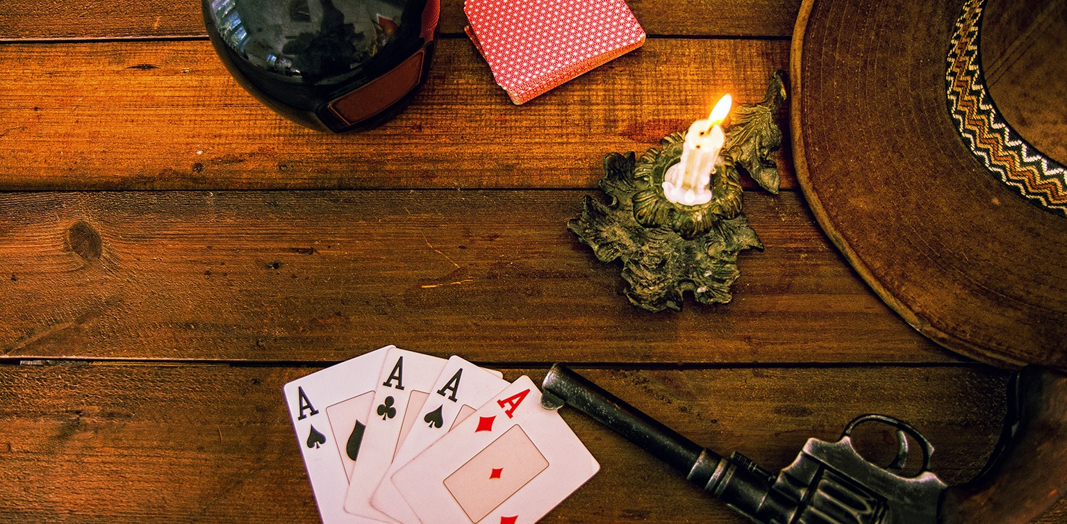 A photo of four aces, a hand gun, candle, Stetson and bottle of whiskey on a wooden table