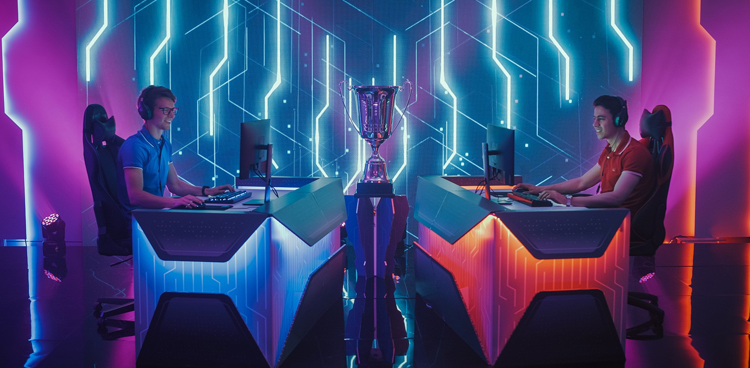 A photographic image of two gamers facing off against each other, with a trophy in-between, in a neon lit room