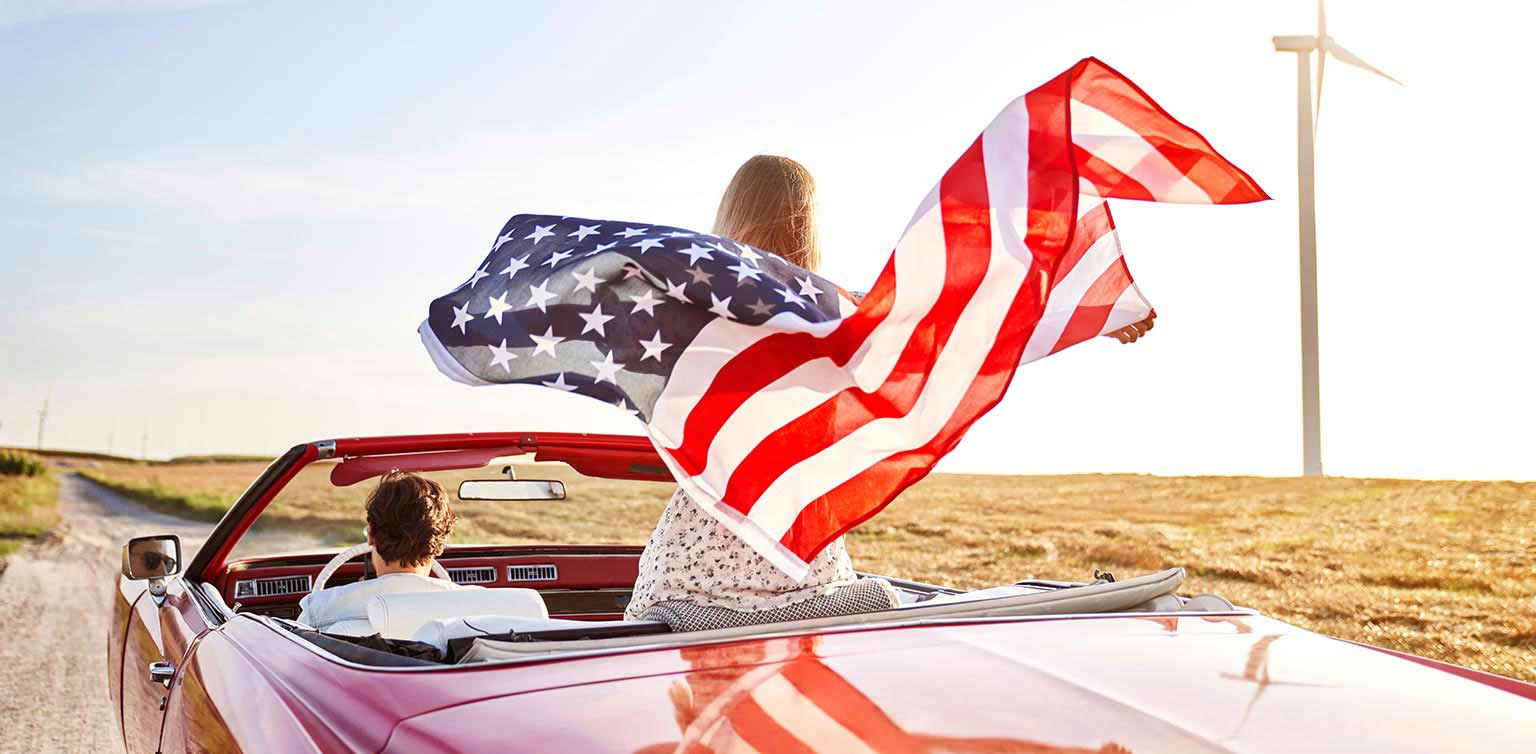 A photo of a man and woman on a road trip in a red convertible with the woman holding the US flag