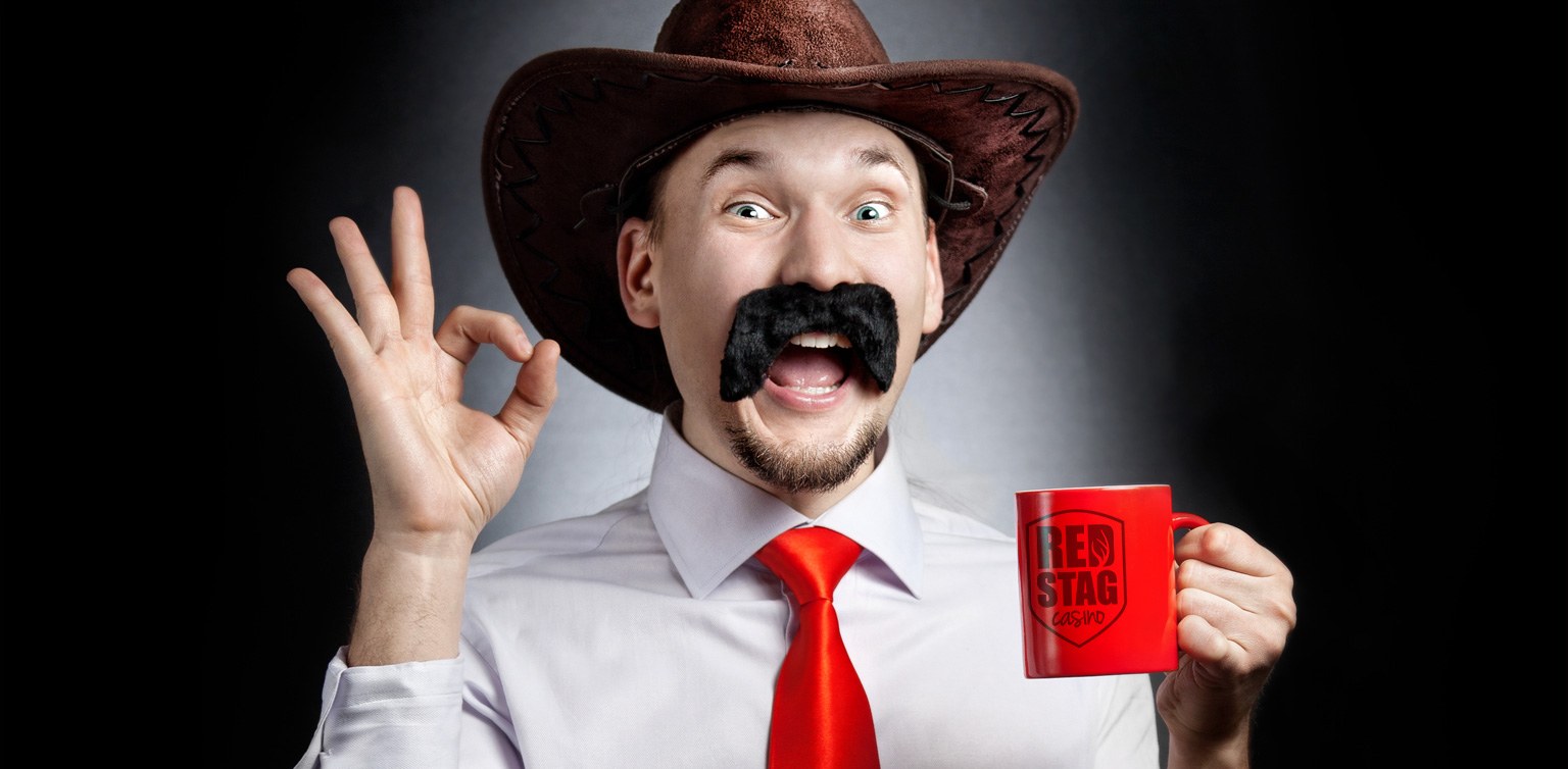 A photograph of a happy guy with a false moustache and Stetson holding a red mug signing OK with his hands on a dark background