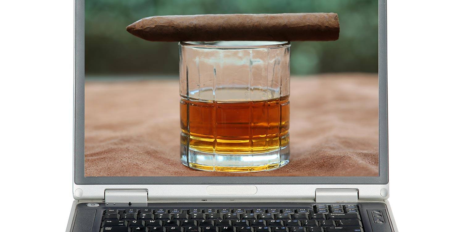A photo of an open laptop displaying a glass of whisky with a cigar balancing on top on the screen